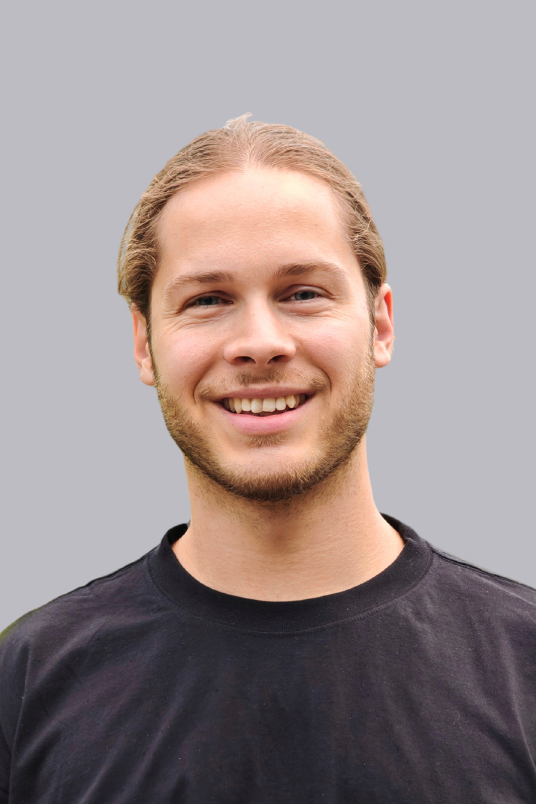 Junger Mann mit hellbraunem Haar, Bart und einem schwarzen T-Shirt, lächelt vor einem grauen Hintergrund.