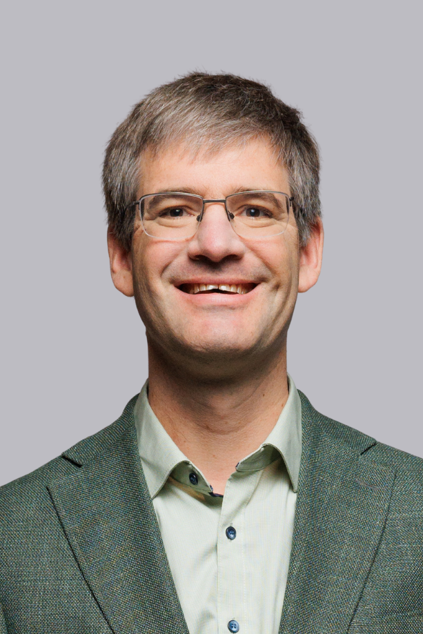 Ein Mann mit Brille, kariertem Anzug und Krawatte lächelt vor hellem grauem Hintergrund.