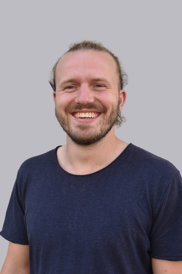 Lächelnder Mann mit langen Haaren in einem blauen T-Shirt vor grauem Hintergrund.