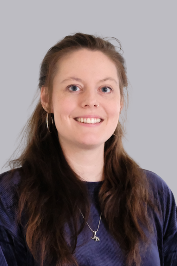 Junge Frau mit langen braunen Haaren, Nase Piercing, Ohrring, im blauen Pullover, lächelnd vor neutralem Hintergrund.
